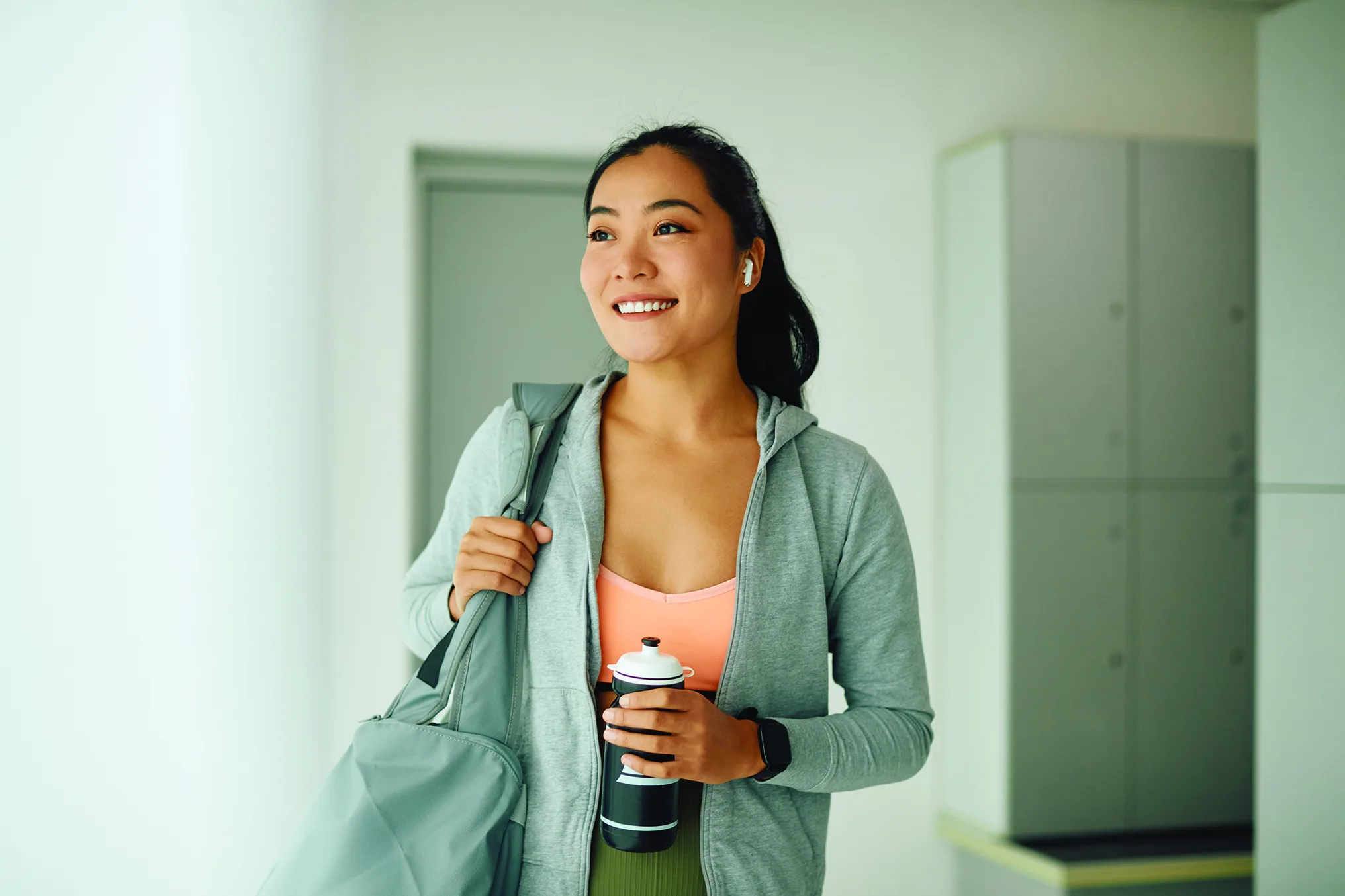 Woman smiling in an empty locker room, she is wearing an athletic top with a sweater unzipped where you can see her olive-green athletic leggings. She is wearing an apple watch and holding her water bottle with her left arm while her right arm braces her gym bag on her right shoulder. She isn't looking at the camera but possible out a window. New pet-friendly apartment homes for rent with private fenced backyards in Kissimmee, FL, near Orlando, FL with pickle ball, dog park, gym, and gated community.