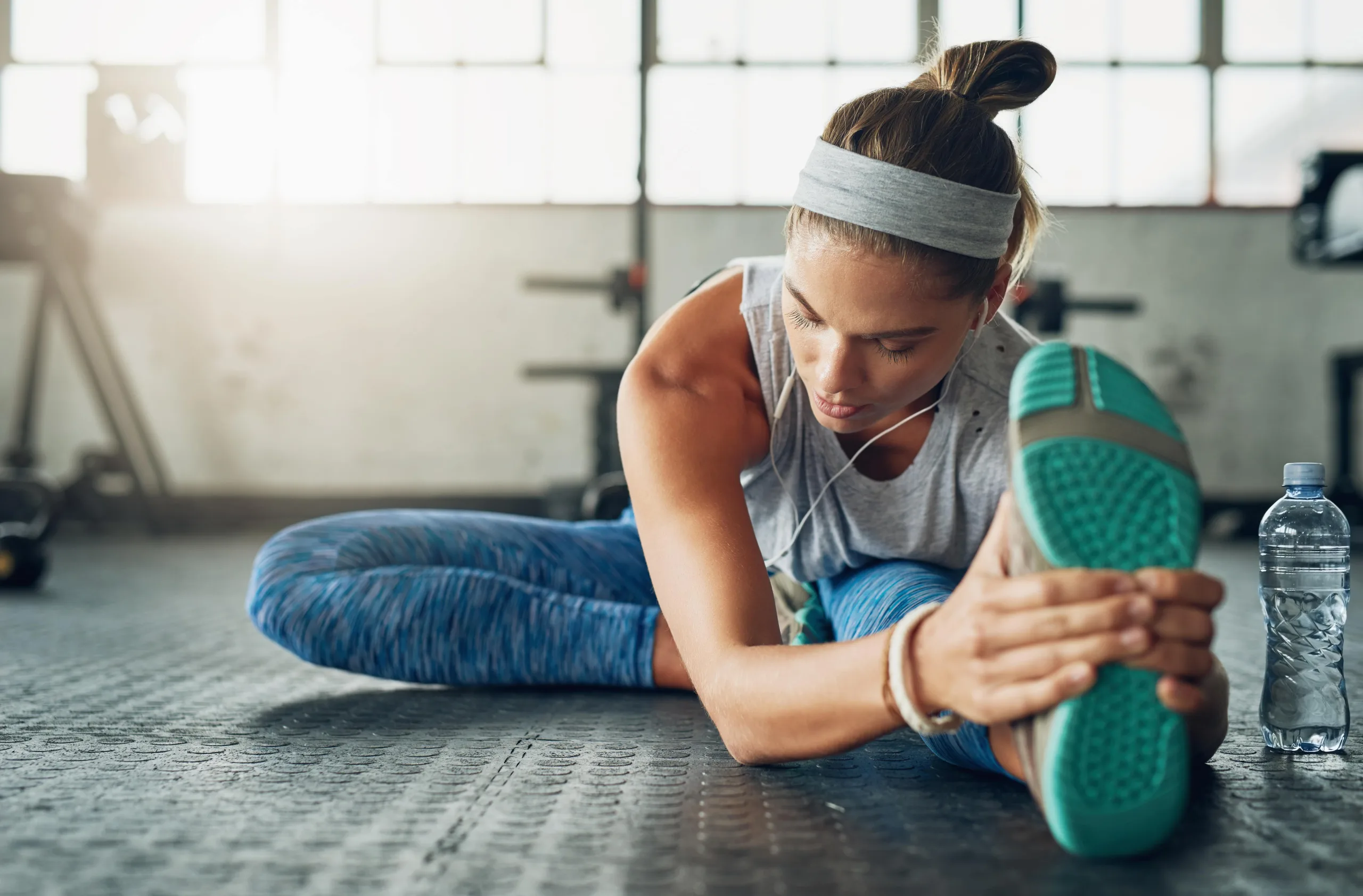 Woman stretching her hamstrings on the gym floor. She has on a grey headband, wire earphones, blue pants and blue soles of her shoes. There is a water bottle beside her, she seems focused and is looking down. New pet-friendly apartment homes for rent with private fenced backyards in Kissimmee, FL, near Orlando, FL with pickle ball, dog park, gym, and gated community.