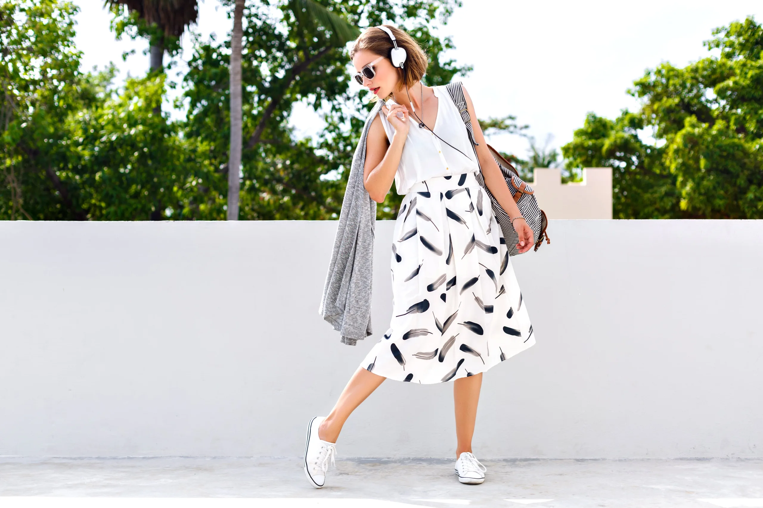 Woman in stylish high waisted skirt and white blouse on a rooftop with tree foliage behind her looking over her shoulder at the ground in headphones, sunglasses, and sneakers. New pet-friendly apartment homes for rent with private fenced backyards in Kissimmee, FL, near Orlando, FL with pickle ball, dog park, gym, and gated community.