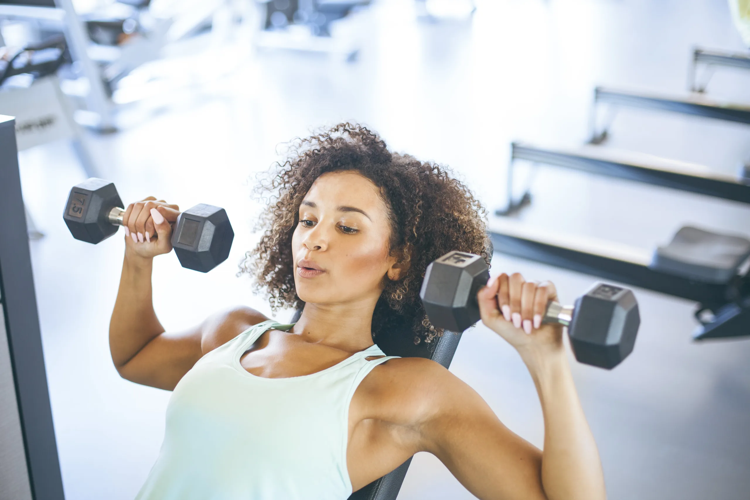 Woman lifting weights as she lays on the bench in a gym. She is wearing a tank top and has curly hair; her lips are pierced as she exhales while exercising. New pet-friendly apartment homes for rent with private fenced backyards in Kissimmee, FL, near Orlando, FL with pickle ball, dog park, gym, and gated community.