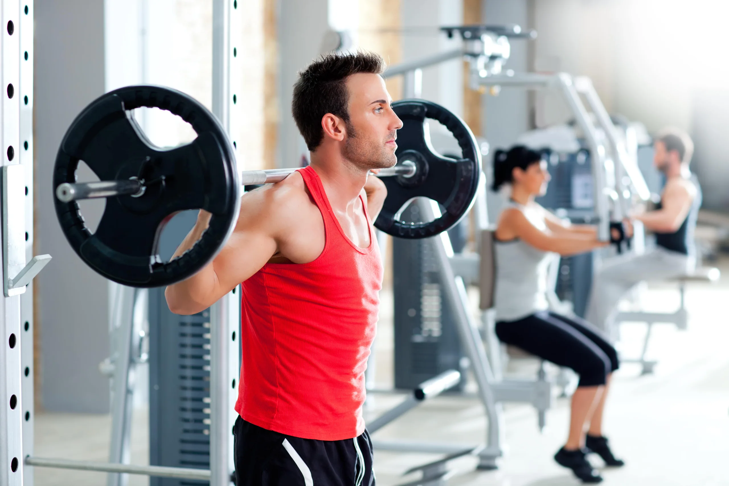 Man in weightroom wearing a red shirt and black shorts with squat bar across his back preparing to squat. In the background you can see a man and a woman on other weight machines and a new clean gym. New pet-friendly apartment homes for rent with private fenced backyards in Kissimmee, FL, near Orlando, FL with pickle ball, dog park, gym, and gated community.