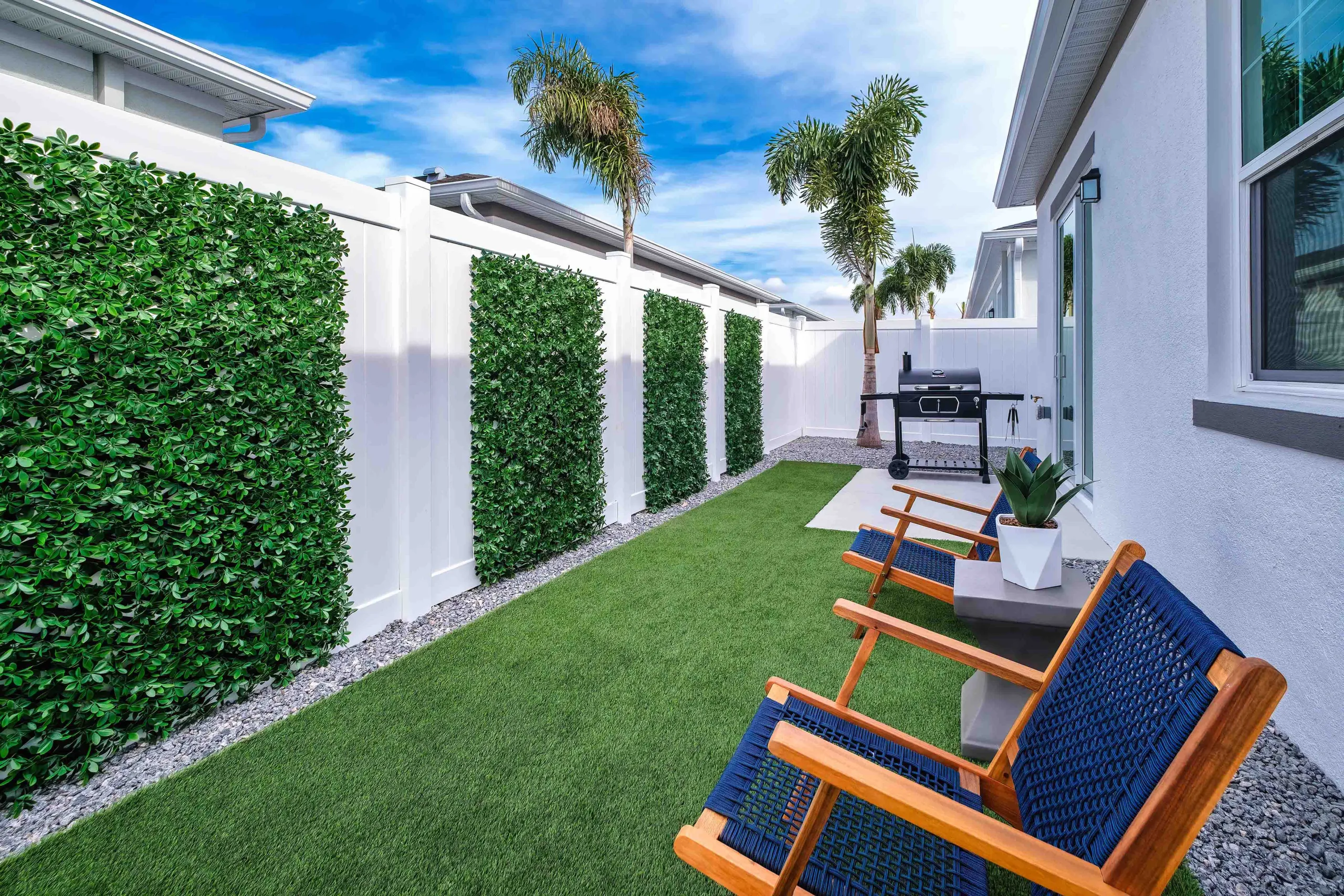 A beautiful Florida backyard scaped with artificial turf, a mature palm tree, seating, a grill, a patio, and a white fence on a beautiful breezy sunny day. New pet-friendly apartment homes for rent with private fenced backyards in Kissimmee, FL, near Orlando, FL with pickle ball, dog park, gym, and gated community.