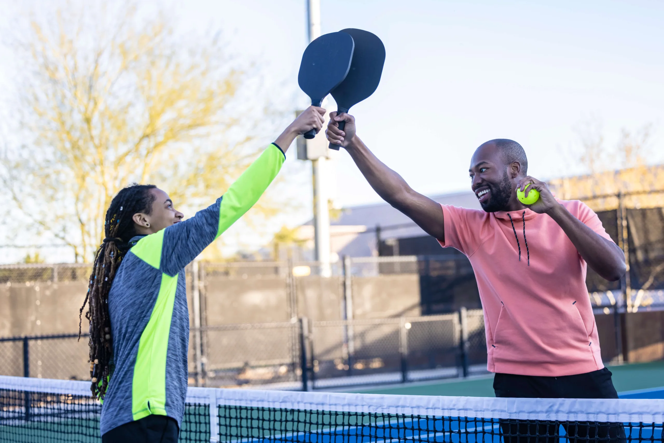 A man and woman happily touch paddles over the pickleball court fence, and the man is holding the pickleball looking at the woman smiling as the woman looks up at the paddles. New pet-friendly apartment homes for rent with private fenced backyards in Kissimmee, FL, near Orlando, FL with pickle ball, dog park, gym, and gated community.